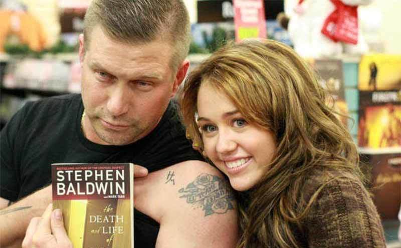 Stephen Baldwin and Miley Cyrus posing together with his book while he points to the initials H and M tattooed on his arm 