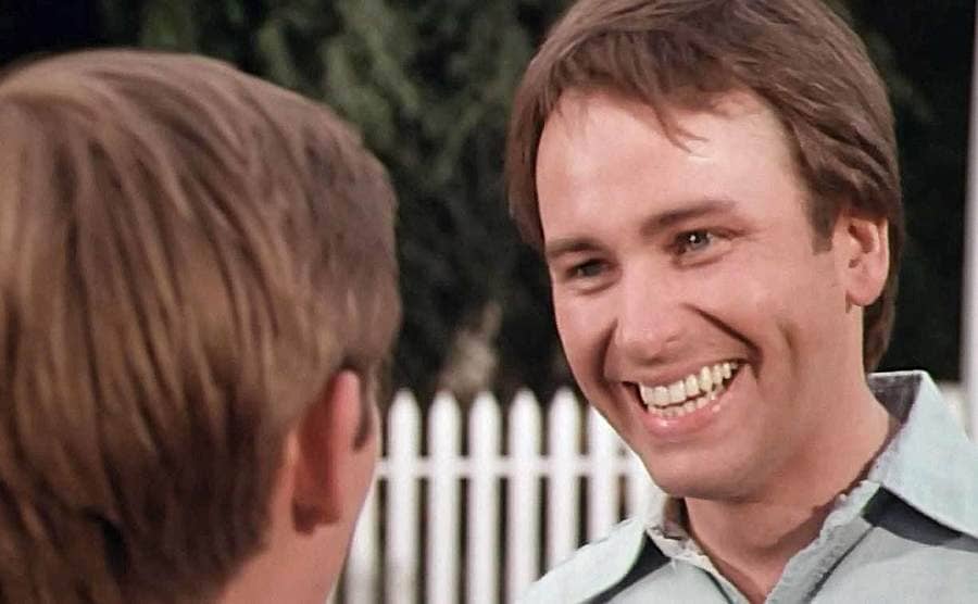 John Ritter in the tv show The Waltons talking to someone in front of a white picket fence 