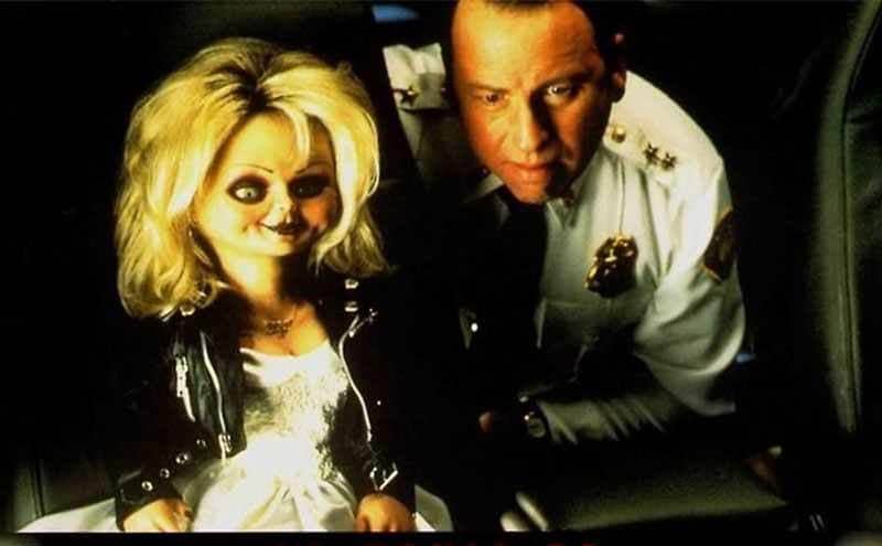 John Ritter dressed as an officer next to a doll from the film Bride of Chucky