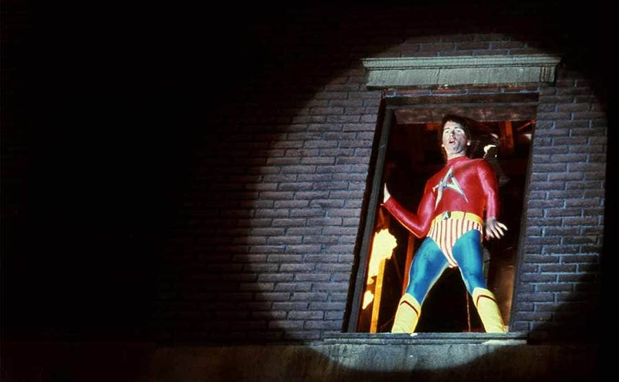 John Ritter as the superhero standing in an open window with a spotlight on him in Hero At Large