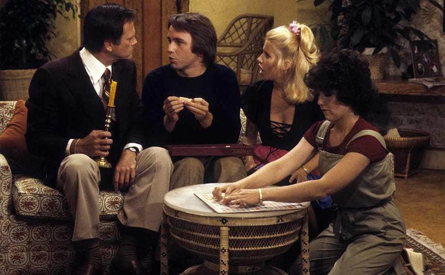 Dick Sargent, John Ritter, Suzanne Somers, and Joyce DeWitt sitting around the couch in an episode of Three’s Company 