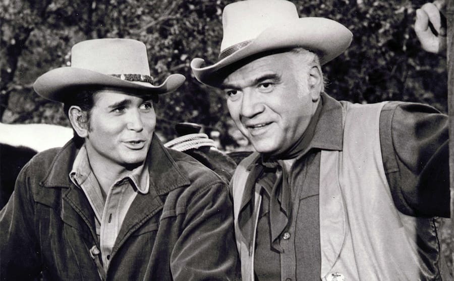 Michael Landon and Lorne Greene posing behind a large wooden fence on the show Bonanza 