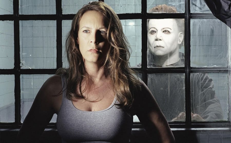 Jamie Lee Curtis standing with her back to a window with a man wearing a mask 
