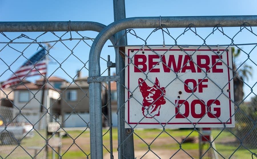 A Beware of Dog sign attached to a fence in front of a blurred out home with a flag 