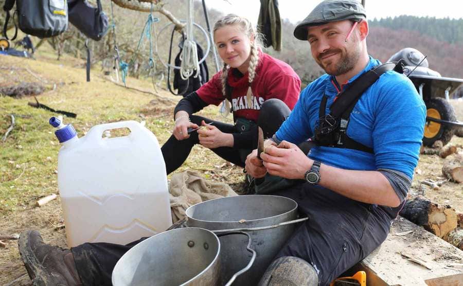 Two contestants peeling potatoes in front of a large jerrycan of water and two large pots 