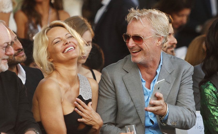 Pamela Anderson and Eddie Irvine laughing together in the front row of a fashion show 