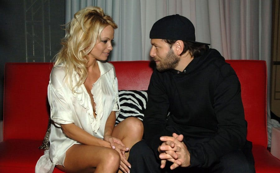 Pamela Anderson and Rick Salomon sitting and talking on a red couch at a party 