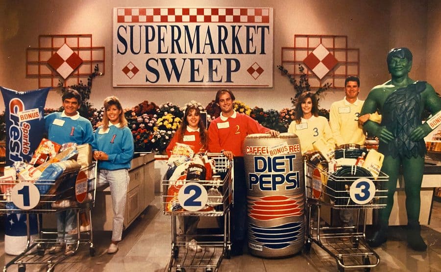 Contestants on Supermarket Sweep with their full carts and large blow-up products standing by the registers 