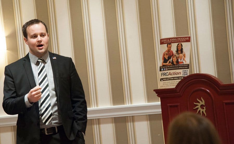 Josh Duggar speaking before a room of people in a suit and tie. 
