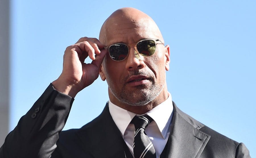 Dwayne Johnson holding onto his sunglasses walking on a sunny day 