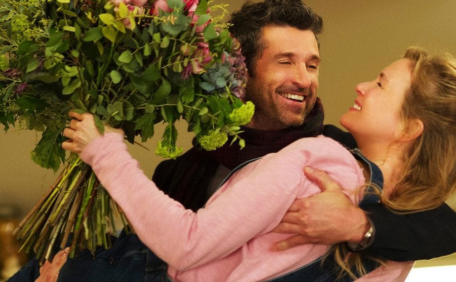 Patrick Dempsey swooping Renee Zellweger off of her feet, who is holding a large bouquet of flowers 