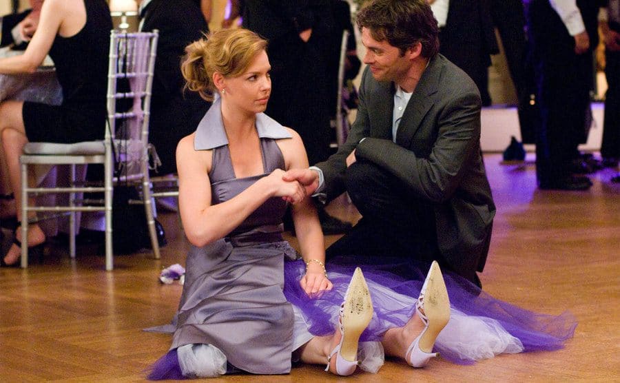 Katherine Heigls sitting on the ground in a purple bridesmaids dress with James Marsden knelling besides her taking her hand to help her up 