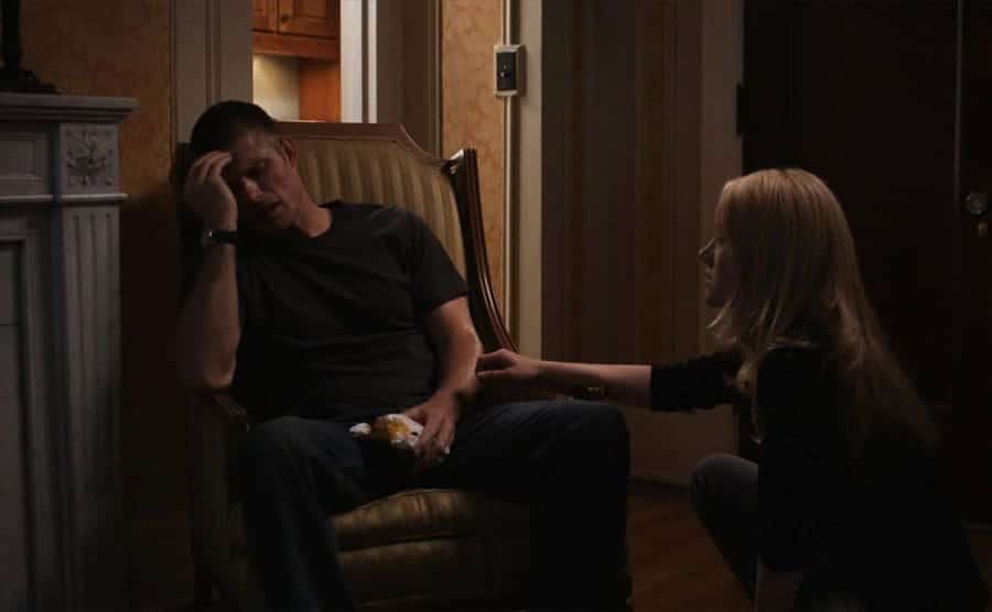 Chris Carmack leaning on his hand in frustration sitting by a fireplace with Rachel Miner consoling him kneeling in front of him 