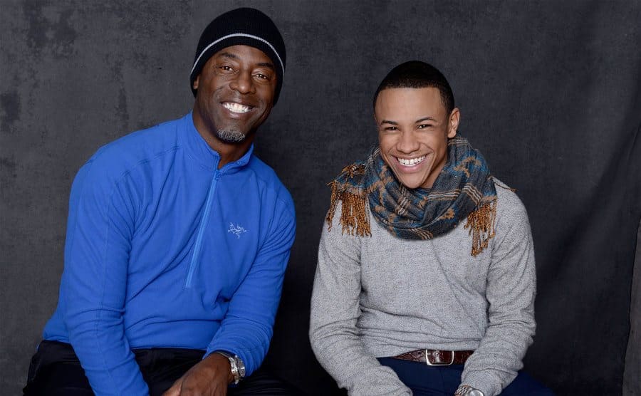 Isaiah Washington and Tequan Richmond posing for a portrait on a wooden bench on front of a grey curtain 