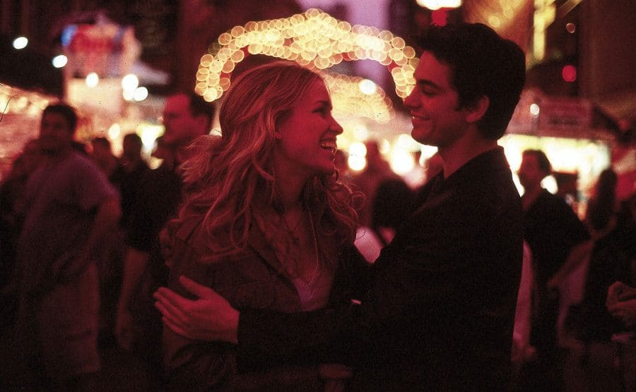 Piper Perabo and Adam Garcia outdoors with places lit up around them 