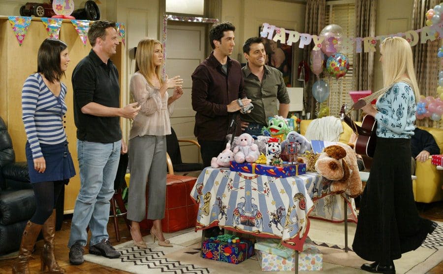 The cast of Friends standing around a table of baby gifts while Phoebe holds a guitar 