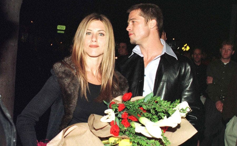 Jennifer Aniston walking with a large bouquet of flowers with Brad Pitt behind her 