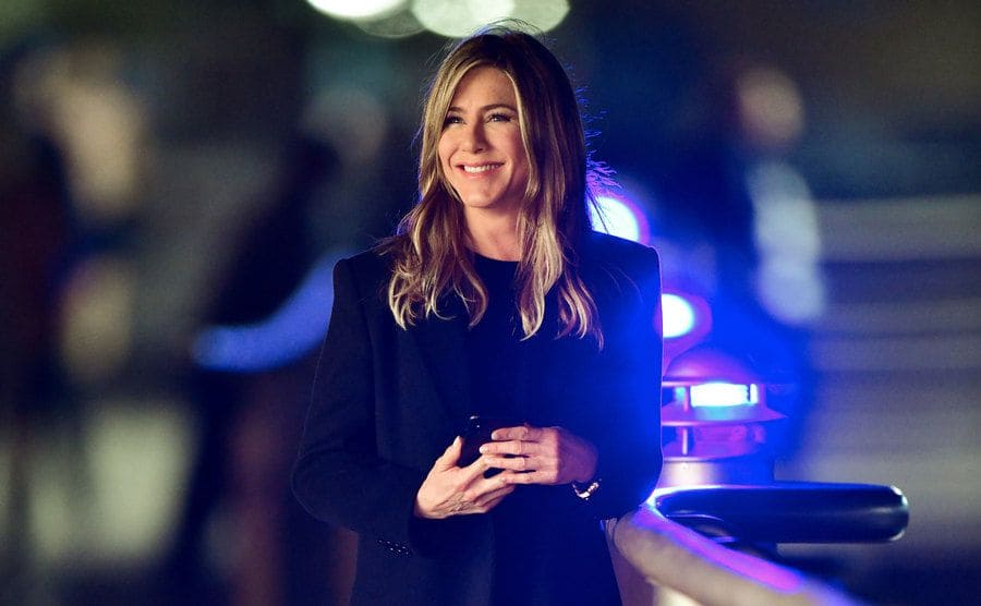 Jennifer Aniston standing outdoors at night with lights blurred around her