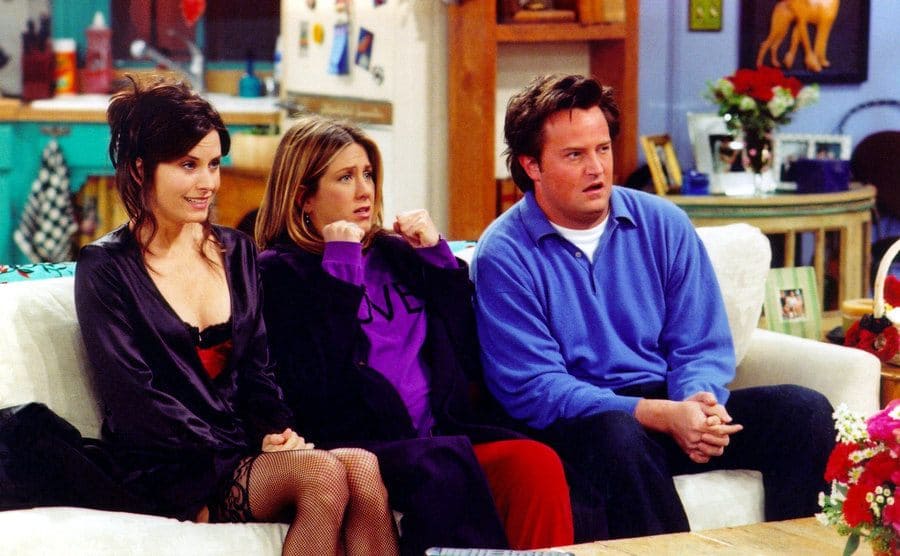 Courteney Cox, Jennifer Aniston, and Matthew Perry sitting on the couch looking scared in a scene from Friends 