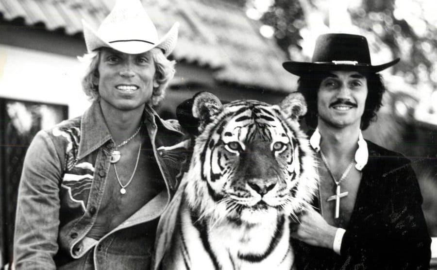 Siegfried and Roy petting a full-grown tiger that sits between them.