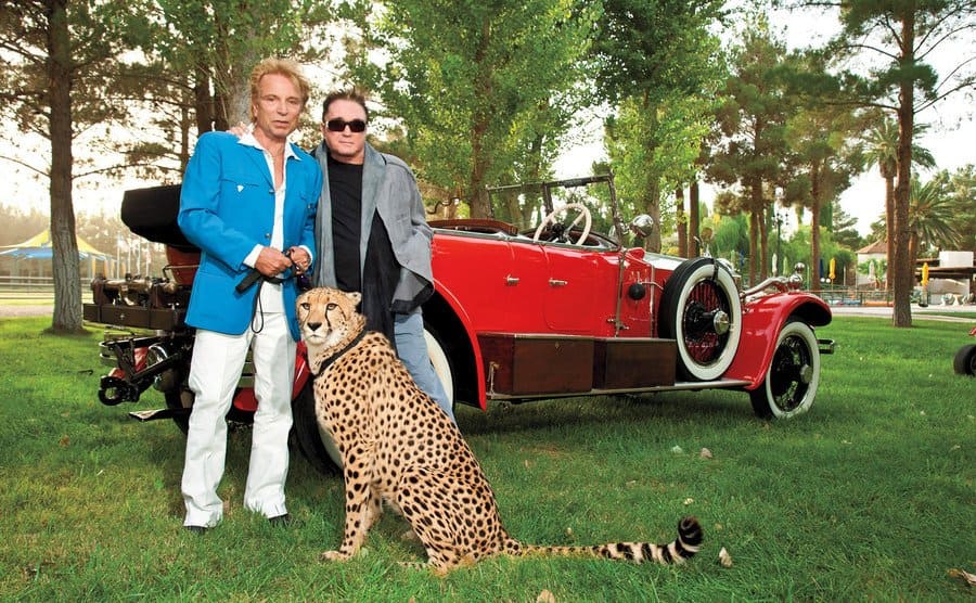 Siegfried and Roy pose with a leopard in front of a vintage red car. 