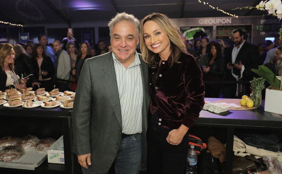 Giada and Lee Schrager posing in front of a bar of plated sandwiches 