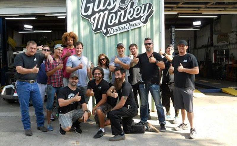 The crew and workers of Gas Monkey Garage are posing for a group picture in front of the sign. 
