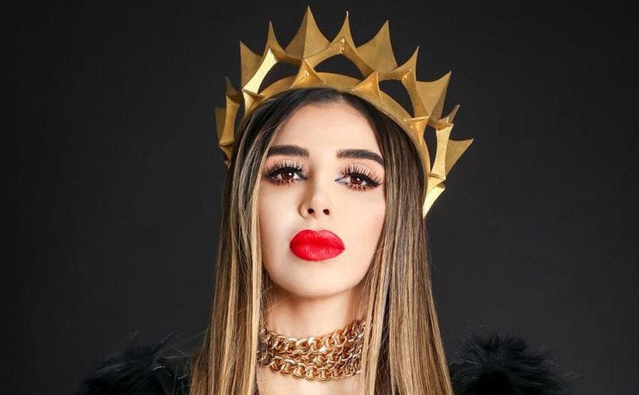 Portrait of Emma Coronel wearing a golden crown standing on a black background.