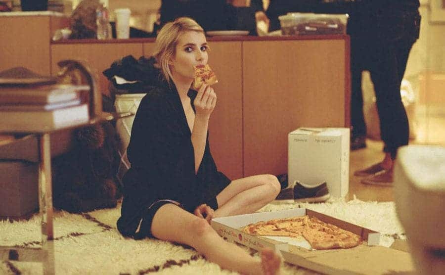 Emma Roberts sits on the floor eating pizza. 