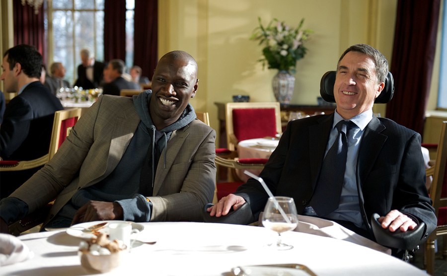 François Cluzet and Omar Sy laugh while sitting at a restaurant. 